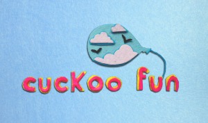 A felt image showing a balloon set against a pale blue felt sky. The balloon has turquoise sky in, with white fluffy clouds and silhoutted birds. Below the balloon, there are letters in pink felt with a yellow felt outline saying "cuckoo fun"